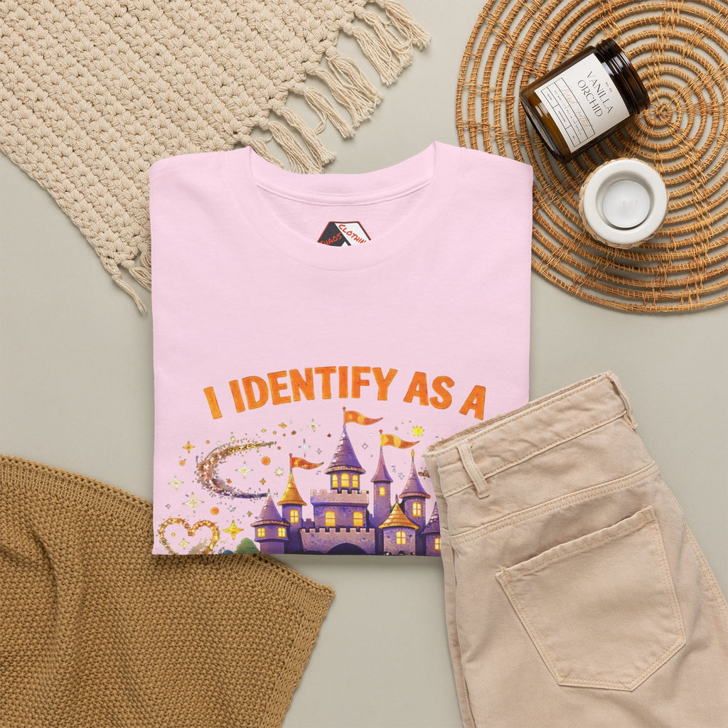 A folded light pink graphic T-shirt is styled in a flat lay with beige pants, a woven blanket, a small candle jar, and a white cup on a neutral surface. The shirt graphic reads 'I IDENTIFY AS A' above a purple castle and colorful sparkly design.