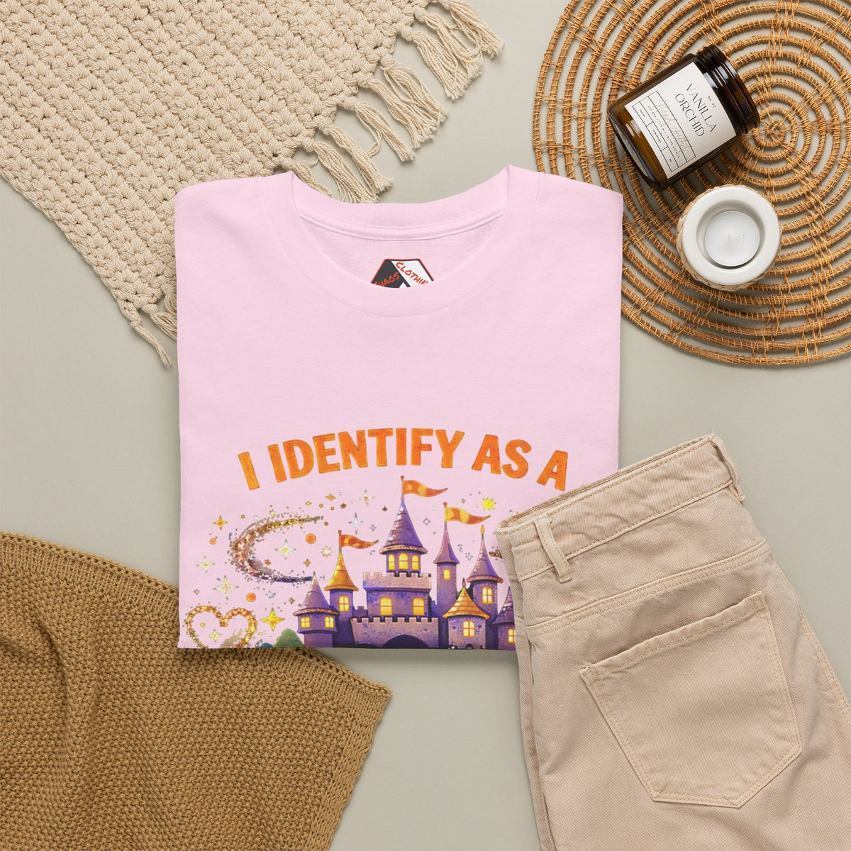 A folded light pink graphic T-shirt is styled in a flat lay with beige pants, a woven blanket, a small candle jar, and a white cup on a neutral surface. The shirt graphic reads 'I IDENTIFY AS A' above a purple castle and colorful sparkly design.