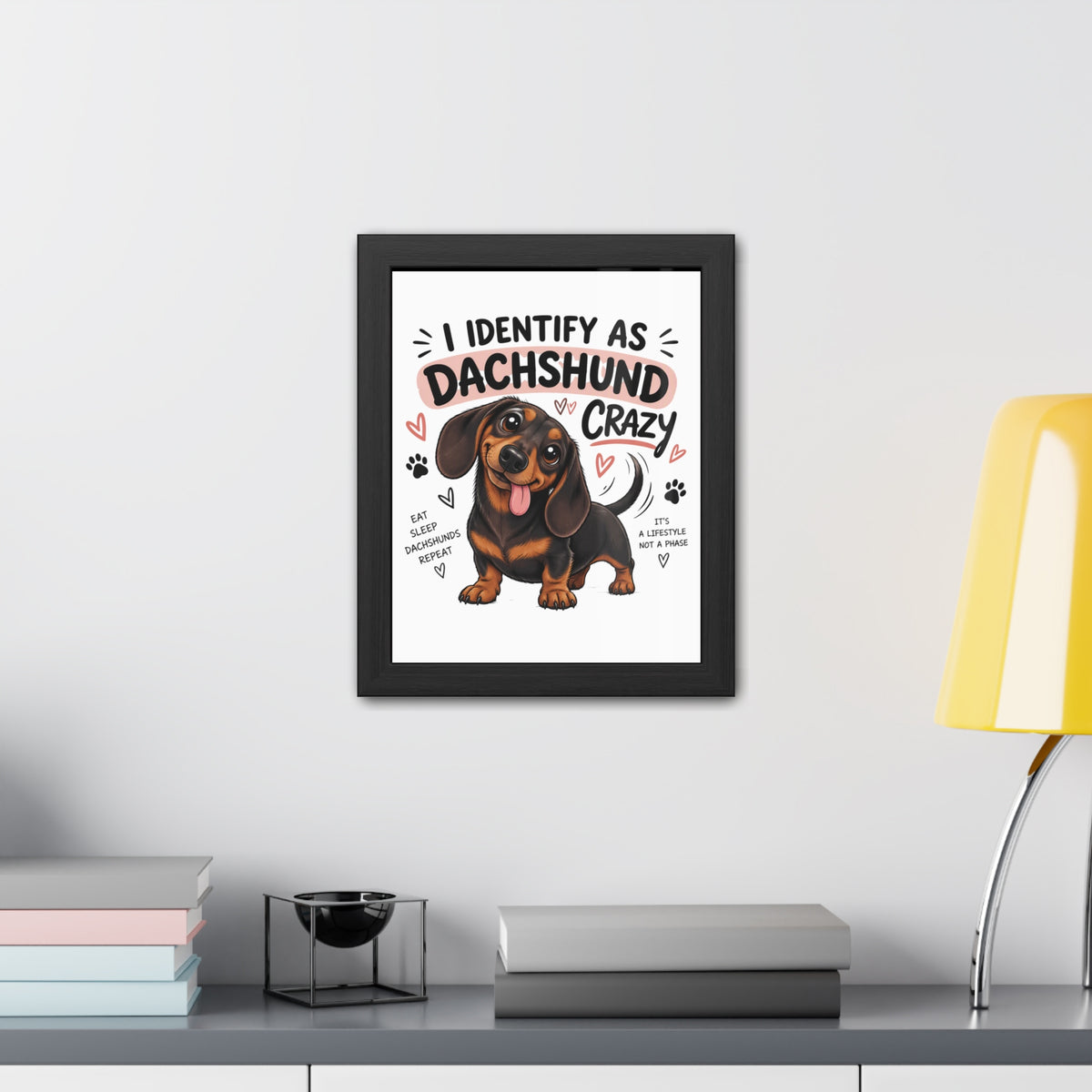 A framed wall art print featuring a cartoon dachshund and the text 'I IDENTIFY AS DACHSHUND CRAZY' is displayed above a shelf with books, a small bowl, and a yellow lamp in a modern room setting.