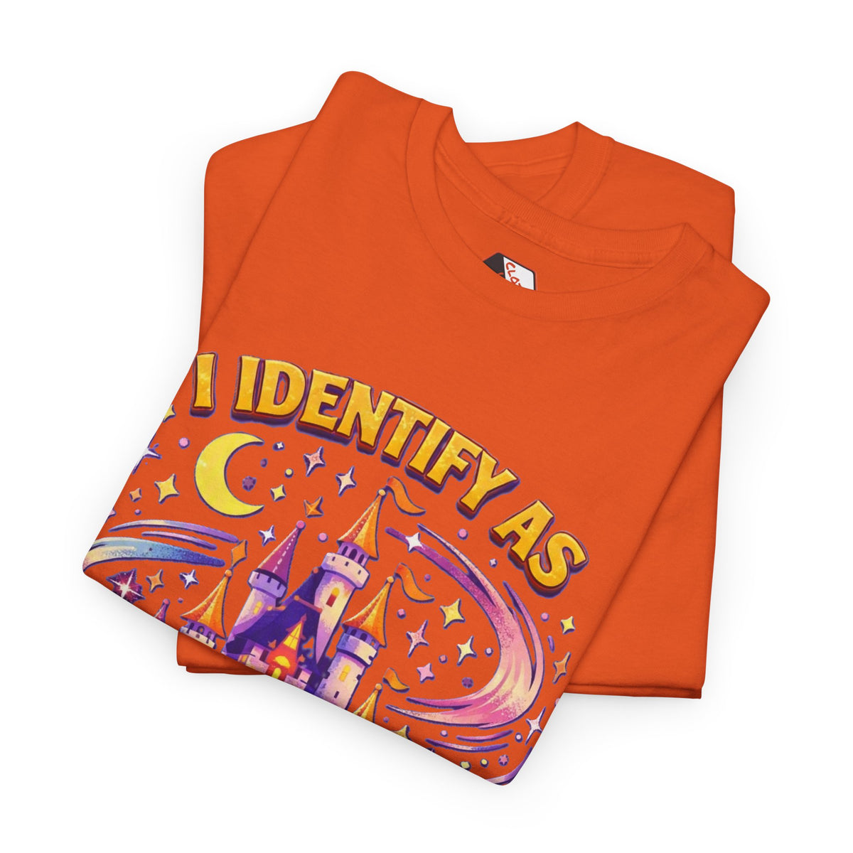 An orange folded t-shirt with a colorful graphic print on the front. The graphic features the text 'I IDENTIFY AS' in yellow with purple outlines, above an illustration of a purple castle, crescent moon, stars, and shooting stars on an orange background.