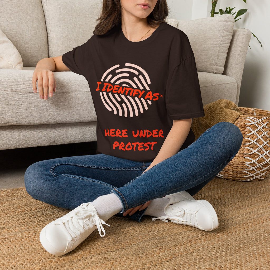 A woman sitting cross-legged on a woven rug in front of a light beige sofa, wearing an oversized dark brown graphic T-shirt with a fingerprint design and the text 'I IDENTIFY AS HERE UNDER PROTEST' in red and white, paired with blue jeans and white sneakers.