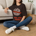 A woman sitting cross-legged on a woven rug in front of a light beige sofa, wearing an oversized dark brown graphic T-shirt with a fingerprint design and the text 'I IDENTIFY AS HERE UNDER PROTEST' in red and white, paired with blue jeans and white sneakers.