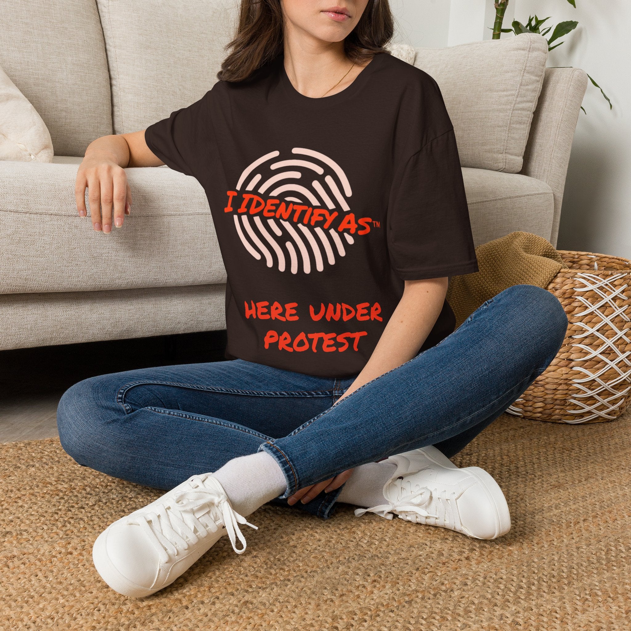 A woman sitting cross-legged on a woven rug in front of a light beige sofa, wearing an oversized dark brown graphic T-shirt with a fingerprint design and the text 'I IDENTIFY AS HERE UNDER PROTEST' in red and white, paired with blue jeans and white sneakers.