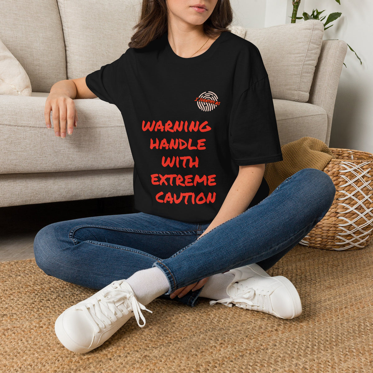 A woman sitting cross-legged on a woven rug in front of a beige sofa, wearing a black oversized T-shirt with a bold orange stacked slogan reading WARNING HANDLE WITH EXTREME CAUTION across the front. A small I Identify As™ circular fingerprint logo sits above the text on the left chest. The design has a chaotic, expressive, personality‑driven feel that matches the brand’s humour‑led style.
