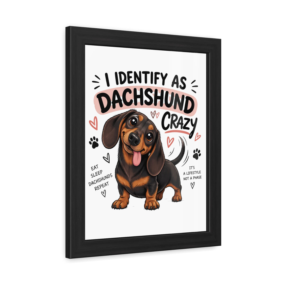 A framed art print featuring a cartoon dachshund with the text 'I IDENTIFY AS DACHSHUND CRAZY' and smaller phrases like 'EAT SLEEP DACHSHUND REPLY' and 'IT'S A LIFESTYLE NOT A PHASE'. The print is shown in a black floating frame against a plain white background.