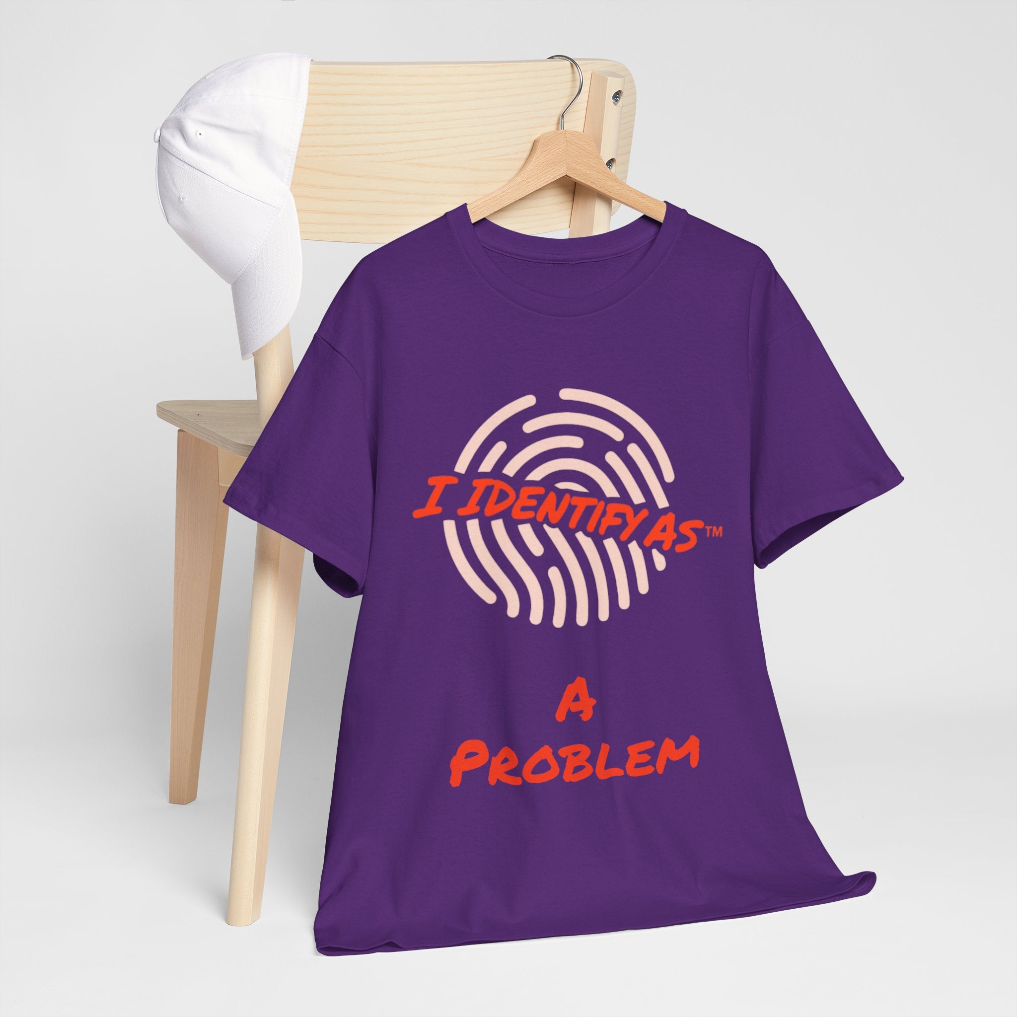 A purple short-sleeve t-shirt on a wooden hanger, styled in front of a light wood chair with a white cap. The shirt features a graphic of a fingerprint in light pink with the text 'I IDENTIFY AS' in orange , and 'A PROBLEM' in orange below.